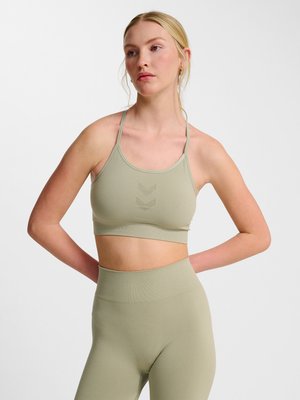 Kvinde med blondt hår står med hænderne bag ryggen iført en lysegrøn sports-bh og matchende højtaljede leggings mod en hvid baggrund.