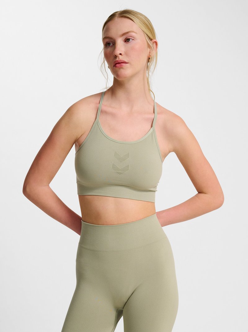 Frau mit blondem Haar steht mit den Händen hinter dem Rücken, trägt einen hellgrünen Sport-BH und passende hoch taillierte Leggings vor weißem Hintergrund.