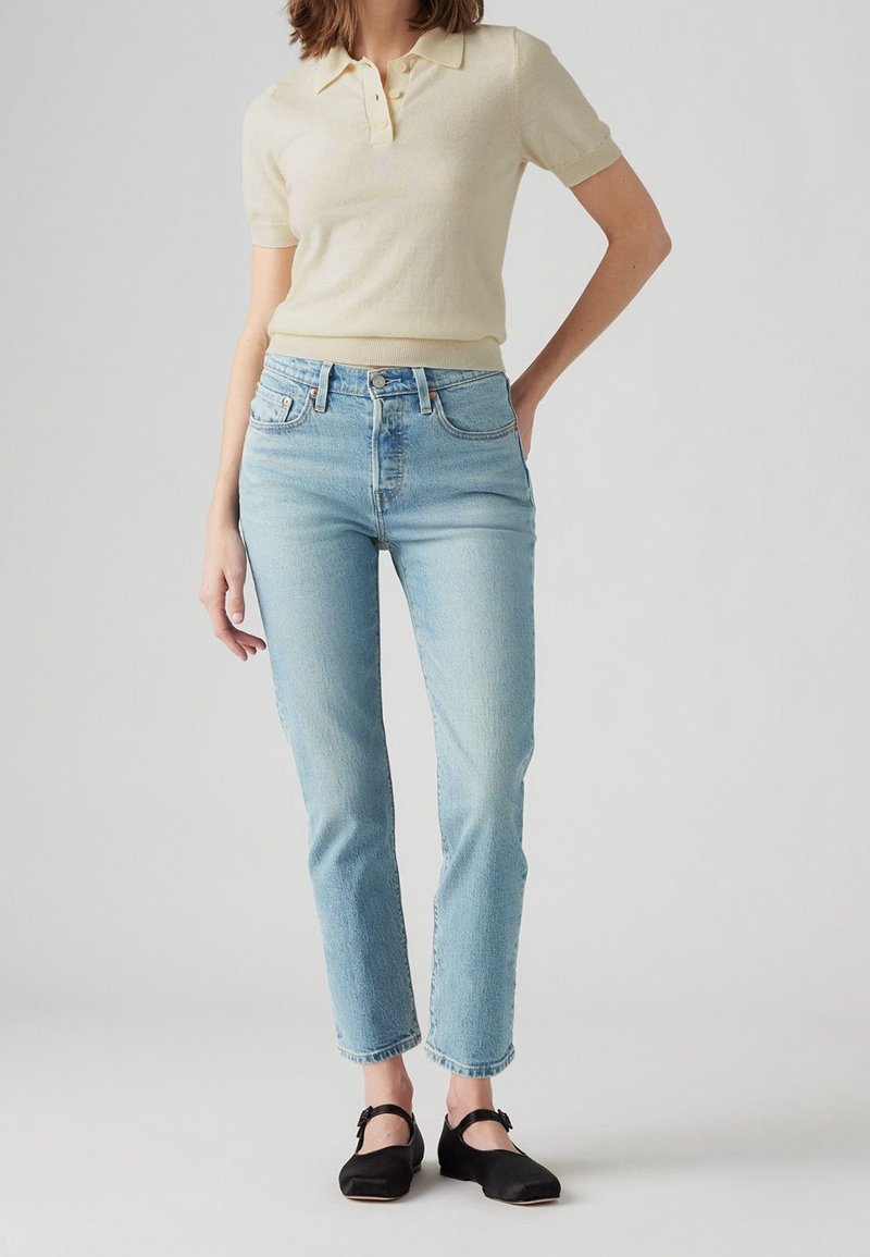 Femme portant un pull polo à manches courtes crème, un jean taille haute bleu clair et des chaussures Mary Jane noires, debout devant un fond uni.