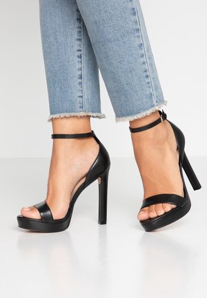 Sorte højhælede sandaler med åben tå, en bred rem over foden og en tynd ankelrem. Glat lædertekstur og platformssål.