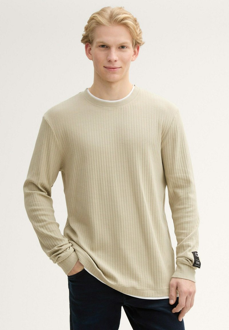 Langarmshirt mit Rundhalsausschnitt in hellem Beige, mit einem strukturierten Waffelmuster, leicht elastisch und einem schwarzen Etikett am linken Ärmel.