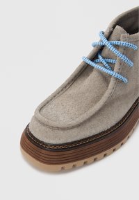 Chaussure en daim gris à surface texturée, avec des lacets bleu à motifs, une semelle brune en couches et une couture détaillée le long du bord.