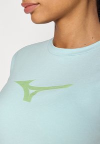 Gaiši zila kokvilnas T-kreklu ar lielu zaļu logotipu dinamiskā dizainā. Apaļš kakls un īsas piedurknes, gluda tekstūra.