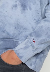 Manica di felpa azzurra tie-dye con logo piccolo ricamato a forma di bandiera, mano parzialmente nella tasca di jeans grigi scuri.