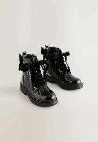 Botas de tobillo de charol negro con cintas de terciopelo y lazos, ojales dorados y suela de goma plana. Cierre lateral con cremallera para facilitar su puesta.