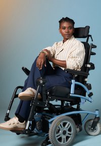 Elektrische rolstoel met blauwe accenten, grote achterwielen en zwarte zitting. Persoon draagt een gestreept shirt en beige sneakers.