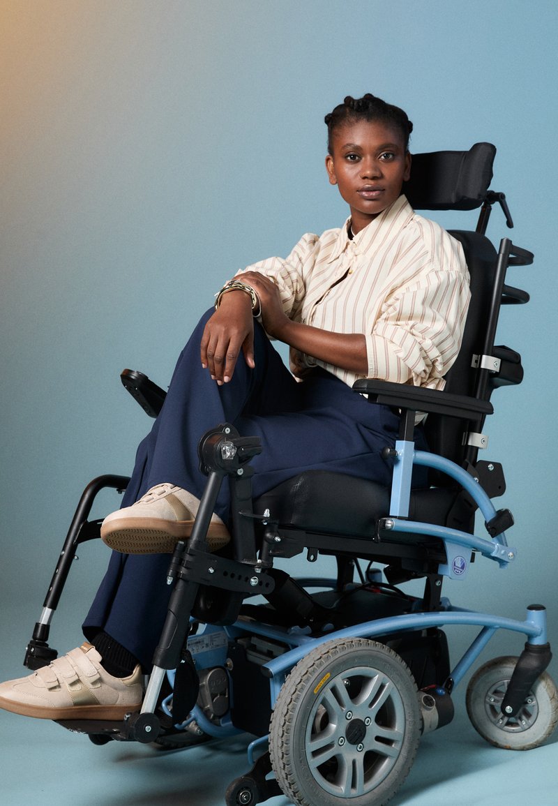 Elektrische rolstoel met blauwe accenten, grote achterwielen en zwarte zitting. Persoon draagt een gestreept shirt en beige sneakers.