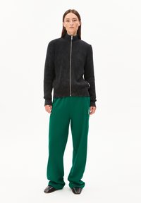 Schwarzer gerippter Pullover mit hohem Kragen und silbernem Reißverschluss, kombiniert mit lockeren grünen Hosen. Die Schuhe sind schwarz mit metallischen Akzenten.