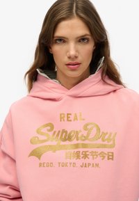 Felpa con cappuccio rosa con testo dorato che dice "REAL SuperDry", caratterizzata da una fodera interna a righe. Tessuto morbido con una vestibilità ampia.