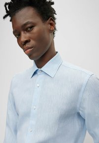 Camicia button-up azzurro chiaro, con una texture e colletto classico. Presenta sottili strisce orizzontali e piccoli bottoni bianchi lungo la parte anteriore.