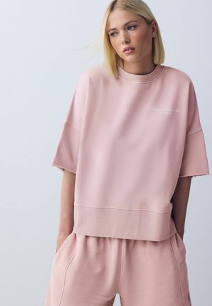 Giovane donna con capelli biondi corti, indossa una felpa larga rosa chiaro e pantaloncini abbinati, mani in tasca, guarda leggermente di lato.