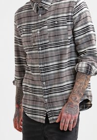 Camicia a quadri grigia e nera con due tasche frontali, maniche arrotolate e un tessuto morbido e texture. Presenta un design del colletto classico.