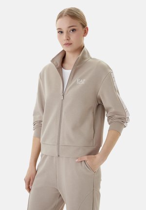 Jeune femme portant une veste zippée beige EA7 et un pantalon assorti, debout avec une main dans la poche sur fond blanc.
