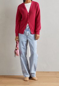 Růžový žebrovaný cardigan s výstřihem do V a knoflíky, kombinovaný se svetle modrými rovnými džíny a hnědými pletenými espadrilkami. Malá růžová kabelka se šňůrkami.