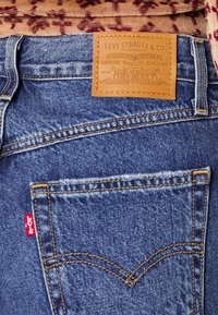 Blauwe denim jeans met een tan leren patch met reliëfprint, opvallende stikpatronen en een klein rood logo op de achterzak.