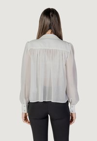 Blusa bianca semi-trasparente con dettaglio a yoke, maniche a sbuffo e polsini con bottoni, presenta un pannello posteriore testurizzato e una parte anteriore liscia.