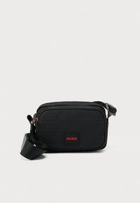 Borsa a tracolla nera in materiale sintetico con finitura testurizzata, caratterizzata da una forma rettangolare, una patch con logo rosso e una tracolla regolabile. Chiusura con cerniera.