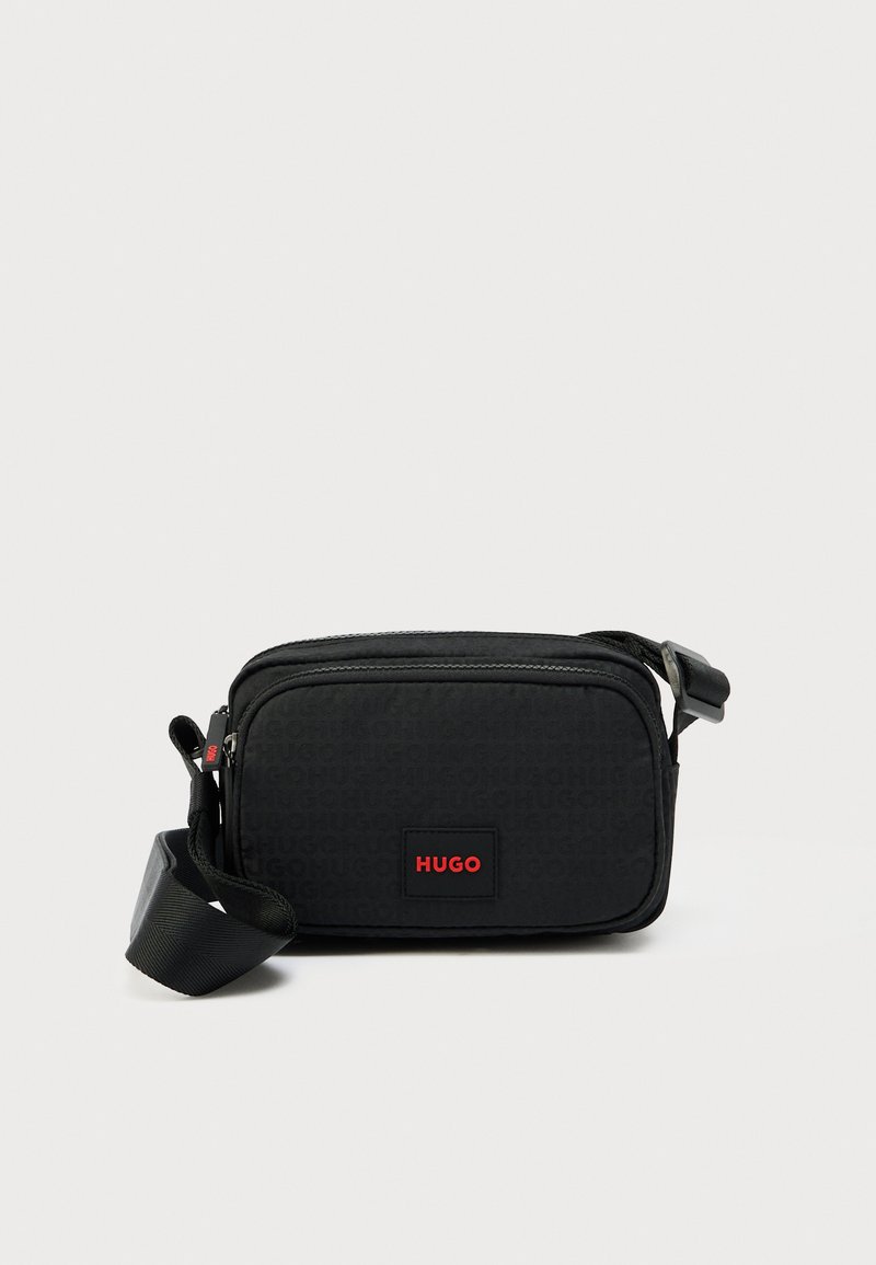 Borsa a tracolla nera in materiale sintetico con finitura testurizzata, caratterizzata da una forma rettangolare, una patch con logo rosso e una tracolla regolabile. Chiusura con cerniera.