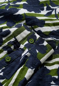 Chemise à manches courtes en tissu tricoté, présentant des motifs floraux bleu marine et verts sur un fond rayé blanc. Comprend des boutons verts.