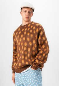 Jonge man in een bruine pluizige trui met oranje vlekken, lichtblauwe jeans met witte stippen, en een lichtgekleurde pet, poseert voor een witte muur.