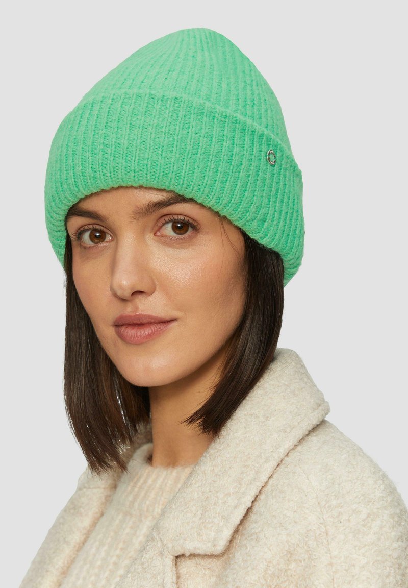 Groene geribbelde gebreide beanie met een omgeslagen rand en een klein metallic accent. Gedragen met een beige gestructureerde jas en een lichte trui.