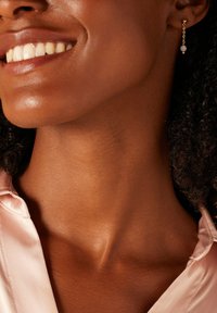 Boucles d'oreilles en chaîne dorée avec une perle centrale, mettant en avant un design simple. Une peau lisse et une chemise en tissu rose clair sont visibles.