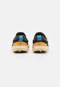 Une paire de chaussures de sport noires et jaunes vue de l'arrière avec des semelles beiges et des tirettes bleues sur un fond blanc.