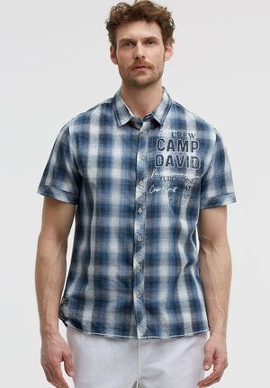 Mann trägt kurzärmeliges blau-weiß kariertes Hemd mit Knöpfen mit Text "Crew Camp David" und weiße Hose vor einfarbigem Hintergrund.