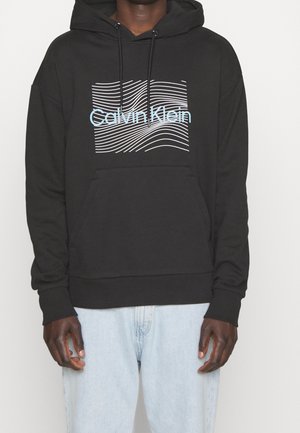 Sudadera negra con líneas onduladas blancas y texto "Calvin Klein" en azul claro, usada por una persona con vaqueros azul claro, manos relajadas a los lados.