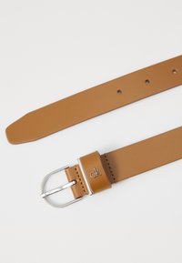 Ceinture en cuir tan avec une boucle en métal argenté, présentant un détail de logo et plusieurs trous pour un ajustement de taille. Texture lisse, forme rectangulaire.
