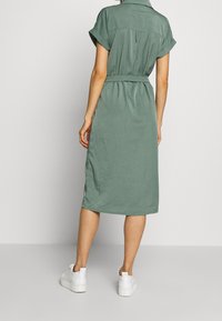 Donna in piedi di spalle che indossa un vestito midi verde con cintura e sneakers bianche, su uno sfondo neutro.