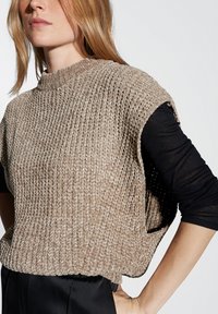 Top senza maniche di colore beige in maglia, con un collo alto, motivo testurizzato e lati aperti, indossato sopra una camicia nera a maniche lunghe.
