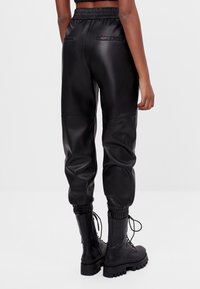 Person klädd i svarta faux läder joggerbyxor med elastiska manschetter och svarta kraftiga snörstövlar, stående mot en enfärgad bakgrund.