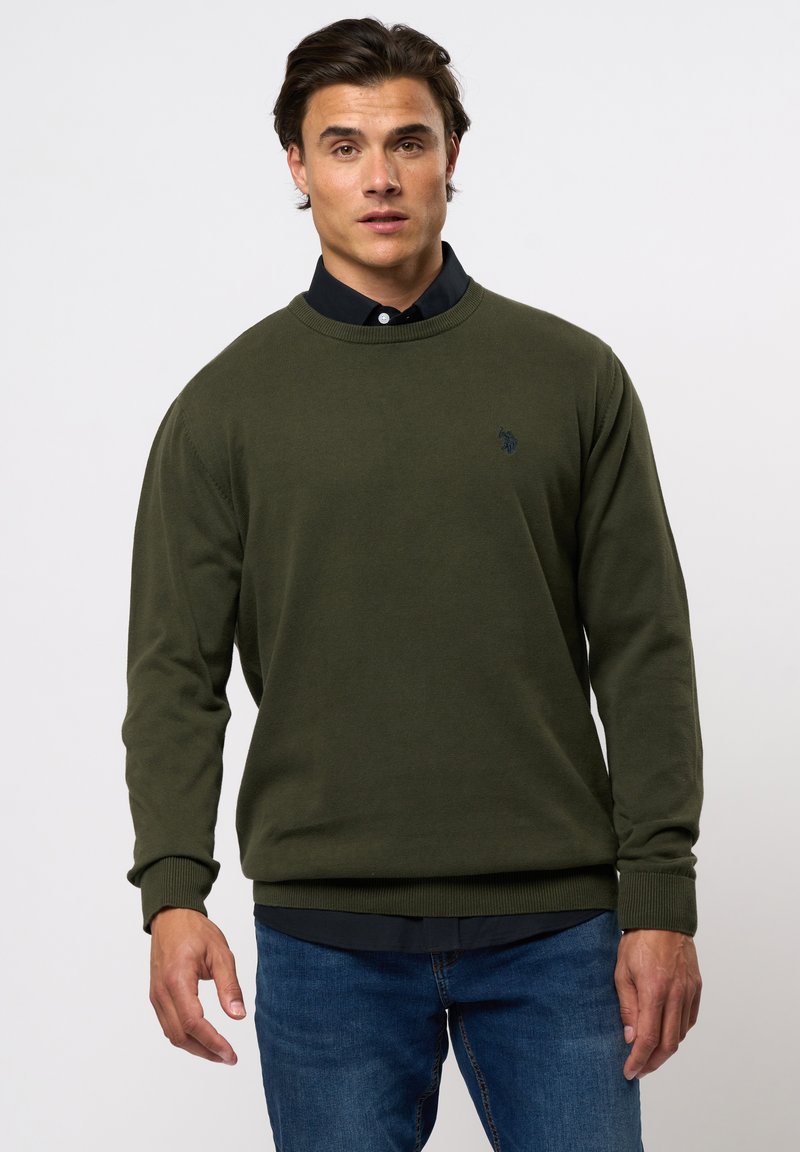 Mørk grønn strikket hettegenser med rund hals, ribbestrikkede mansjetter og en liten marineblå logo på brystet. Bærers over en svart skjorte med krage.