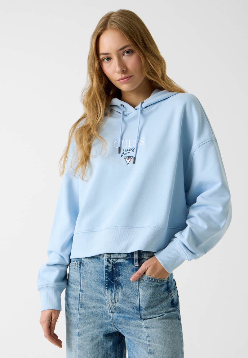 Sweat à capuche court bleu clair avec cordon de serrage, arborant un logo brodé blanc et un écusson triangulaire. Associé à un jean en denim délavé taille haute.
