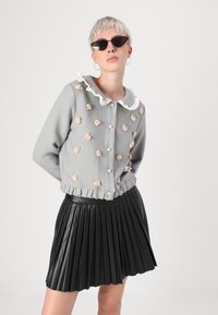 Cardigan gris texturé avec des embellissements floraux, un col rufflé blanc et des boutons en perles, associé à une jupe en cuir noir plissée.