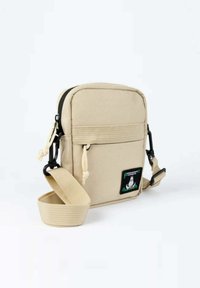 Beige olkalaukku, jossa on teksturoitu kangas, vetoketjusuljenta, säädettävä hihna ja etupuolella sarjakuvahahmon kuvio.