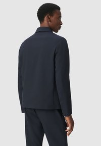 Blazer azul marino oscuro con un corte estructurado, que cuenta con una sola abertura trasera y una tela suave. Exhibido en un modelo desde un ángulo lateral.