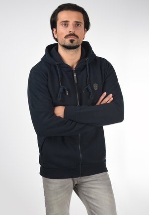 Námořnická modrá mikina na zip s kapucí na stažení, texturovaná tkanina a detail loga na hrudi. V kombinaci se světlými šedými džíny.