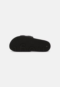 Semelle de sandale noire avec surface texturée, présentant des détails de logo en relief et une forme incurvée. Fabriquée en caoutchouc pour plus de durabilité.