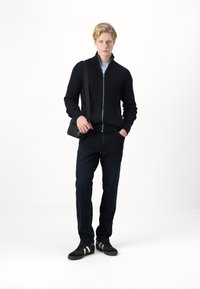Crna zip-up pletena jakna, plava majica ispod, tamne traperice, crna torba za crossbody i crne tenisice s bijelim prugama.