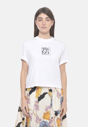 Donna con capelli castano lisci, che indossa una maglietta bianca a maniche corte con logo in scrittura nera e una gonna a fantasia floreale nei colori giallo, rosa e blu.