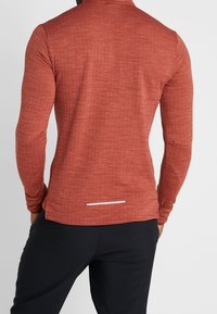 Person in einem enganliegenden, langärmligen rostrot Shirt und schwarzen Hosen, von hinten vor einem schlichten Hintergrund abgebildet.