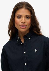 Camisa de botones azul marino con cuello, que presenta botones blancos y un pequeño logo en el lado izquierdo del pecho. Tejido suave, corte relajado.