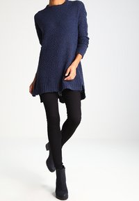 Femme portant un pull long texturé bleu marine, des leggings noirs et des bottines noires, debout devant un fond clair uni.