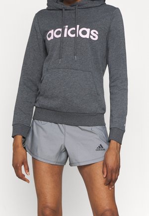 Person iført en mørkegrå hættetrøje med teksten "adidas" og lysegrå adidas-shorts med logo på venstre ben, stående med armene afslappede.