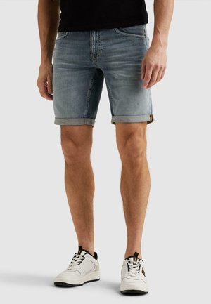 Mand iført lyseblå, opsmøgede denimshorts og hvide sneakers, stående på en ensfarvet baggrund.