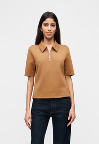 Γυναίκα που φοράει κοντομάνικη πόλο μπλούζα χρώματος καμηλοπάρδαλη με φερμουάρ στο μπροστινό μέρος και σκουρομπλε τζιν, στέκεται μπροστά σε ένα απλό φωτεινό φόντο.