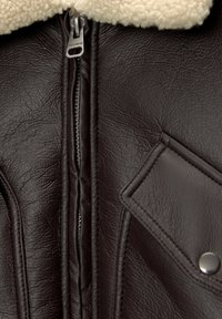 Bruine neplederen jas met een crème schapenvachtkraag. Heeft een zilveren rits en een diamanten vormig zakpatch met een drukknoop.