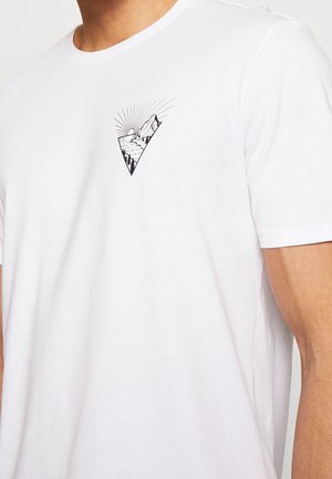 Hvid t-shirt med et lille sort geometrisk bjerg- og solmotiv på venstre brystområde, båret af en person.